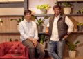 Two startup founders posing in a modern office space with indoor plants and wooden shelving in the background.