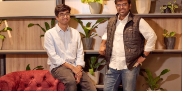Two startup founders posing in a modern office space with indoor plants and wooden shelving in the background.